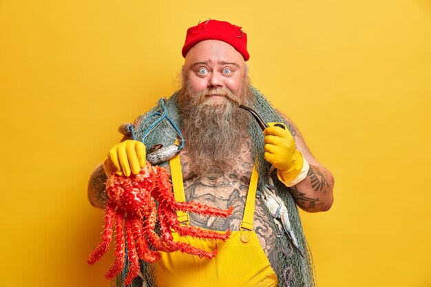 La foto del marinaio paffuto e barbuto ha catturato un grosso polpo rosso che trasporta una rete da pesca vestita con una tuta ha il corpo tatuato