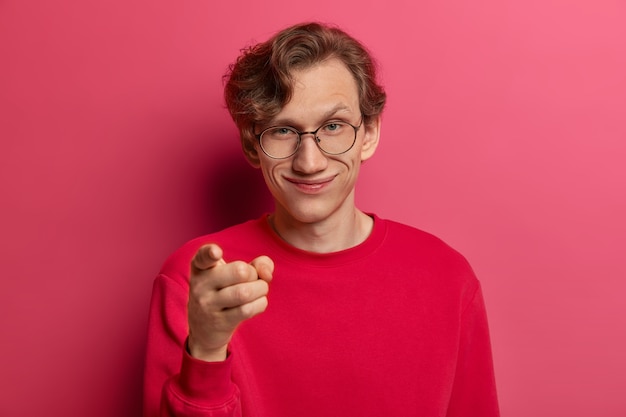 La foto del nerd maschio misterioso felice indica con il dito indice e guarda con curiosità, seleziona qualcuno, indossa occhiali e maglione casual, isolato sul muro rosa. Hmm, interessante
