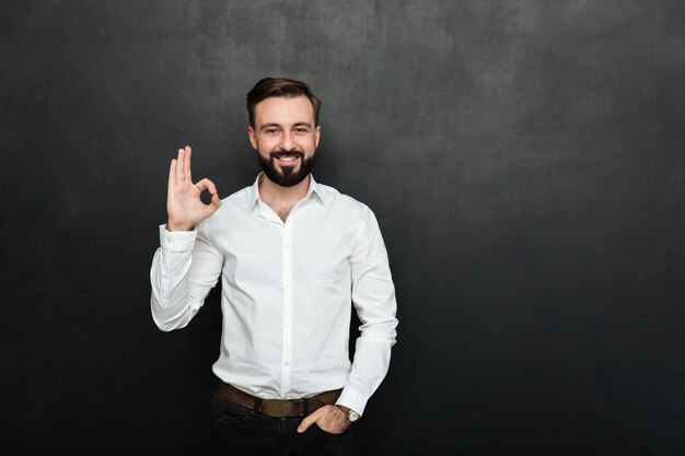 La foto del ragazzo unshaved in ufficio che sorride e che gesturing con il segno GIUSTO che esprime tutto va bene, isolata sopra la grafite