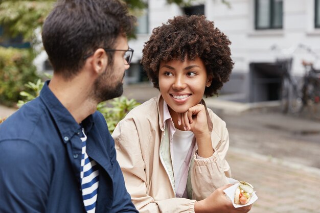 La foto del turista di razza mista gode di una comunicazione vivace, mangia un fast food per strada, è di buon umore.