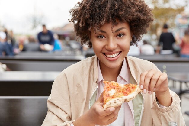 La foto dell'adolescente soddisfatta con la pelle sana e scura, gode di un pasto delizioso, tiene un pezzo di pizza