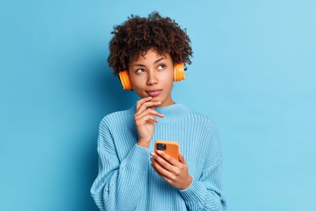 La foto della bella donna afroamericana ascolta la musica con lo smartphone e le cuffie ha un'espressione premurosa vestita di jumpper lavorato a maglia