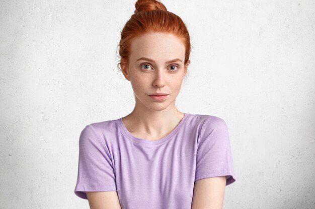 La foto della bella donna dai capelli rossa seria si è vestita in maglietta casuale viola, isolata sopra lo studio bianco