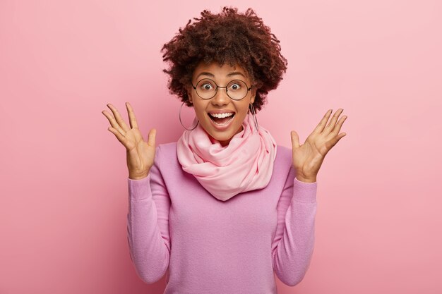 La foto della bella donna felice ed eccitata ha un taglio di capelli afro, alza i palmi, reagisce alle buone notizie, modella su un muro roseo dello studio