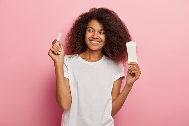 La foto della donna afroamericana felice tiene il tampone e il tovagliolo sanitario, vestita di maglietta bianca, isolata sopra la parete rosa Donne, pms