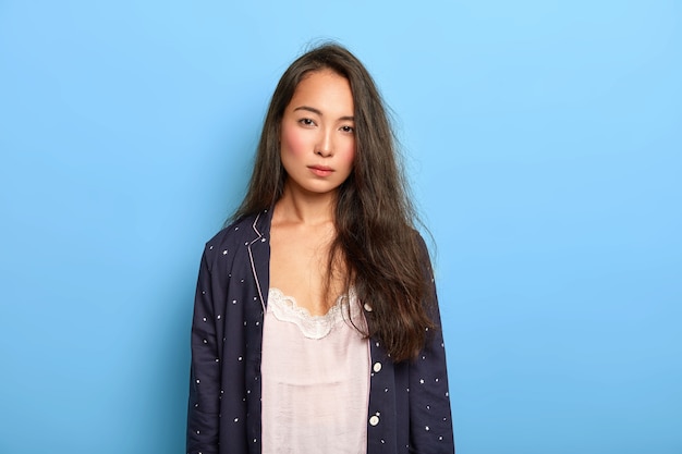 La foto della donna asiatica castana insoddisfatta seria guarda con stanchezza alla macchina fotografica, non ha abbastanza sonno, indossa la camicia da notte