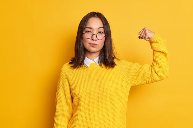 La foto della donna asiatica castana seria alza il braccio e mostra che la sua forza ha muscoli forti sta in piedi interni sicuri di sé indossa un maglione giallo e occhiali ottici rotondi. Concetto di potere delle donne