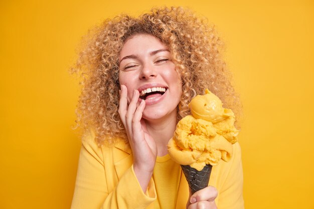 La foto della donna dai capelli ricci felicissima si sente divertita sorrisi tiene ampiamente gli occhi chiusi si diverte a mangiare un delizioso gelato al sapore di mango posa con un delizioso dessert congelato isolato sulla parete gialla.