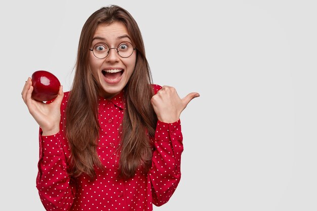 La foto della donna dai capelli scura lieta con un'espressione felicissima, punta sul lato destro, porta una succosa mela rossa