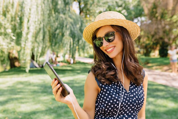 La foto della donna di stile sta camminando nel parco estivo con indosso un cappello estivo e occhiali da sole neri e un vestito carino. Ascolta musica e balla con grandi emozioni.