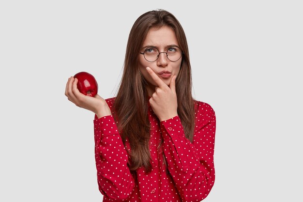 La foto della donna graziosa premurosa seria tiene il mento, porta la mela, ha un'espressione contemplativa, indossa la camicia rossa