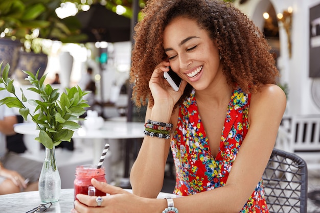 La foto della donna riccia dalla pelle scura positiva indossa una camicetta alla moda, ha una piacevole conversazione telefonica durante il riposo nella caffetteria all'aperto