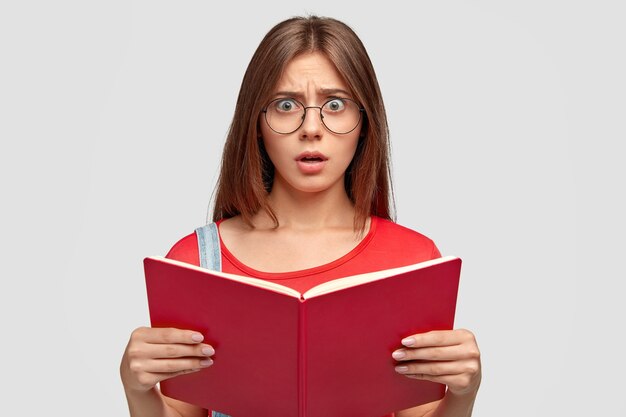 La foto della giovane donna caucasica emotiva stupefatta guarda con stupore, tiene il libro rosso, deve imparare molto per la lezione successiva, indossa occhiali rotondi, isolato su un muro bianco