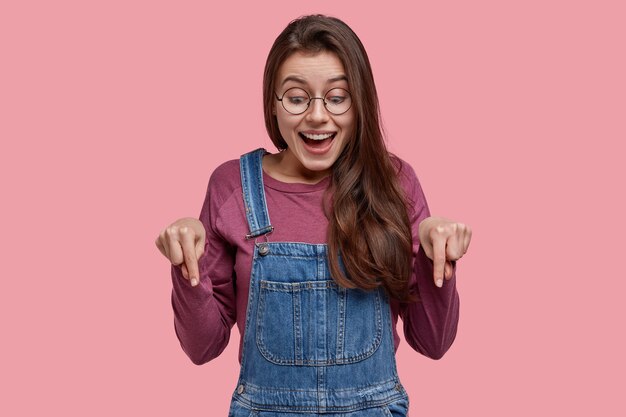 La foto della giovane signora europea allegra attraente con capelli scuri dritti, punta verso il basso, ha un'espressione felice, vestita in abiti casual