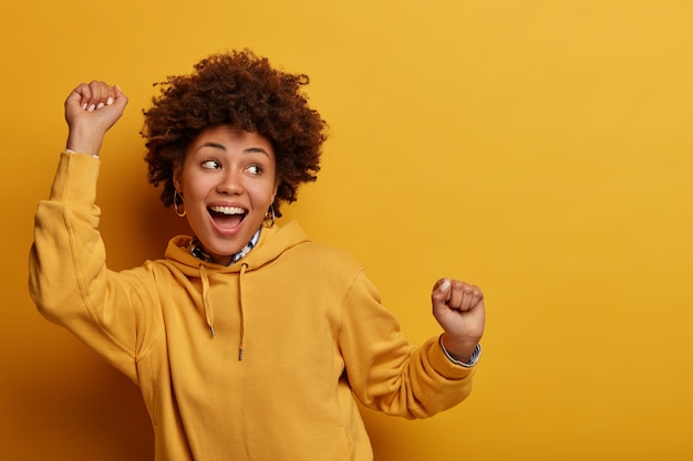 La foto della ragazza afroamericana balla fortunata, alza le mani in segno di evviva, si sente come una campionessa dopo aver ottenuto il trionfo, guarda felicemente da qualche parte, si diverte, sente il ritmo della musica, isolata sul muro giallo