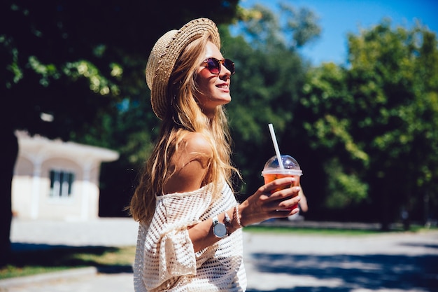 La foto di estate della giovane donna affascinante in occhiali da sole, tiene una tazza di cocktail fresco