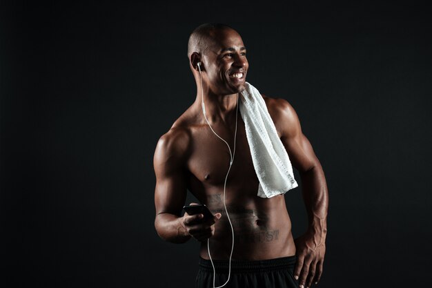 La foto di giovane uomo afroamericano sorridente di sport con il telefono della tenuta dell'asciugamano ed ascolta la musica