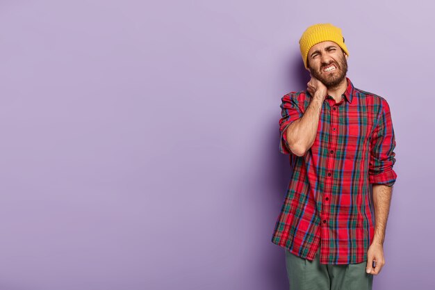 La foto di un giovane insoddisfatto con setole, sente rigidità al collo, inclina la testa, stringe i denti, vestito con una camicia a scacchi, si erge su uno sfondo viola spazio vuoto per il tuo contenuto promozionale