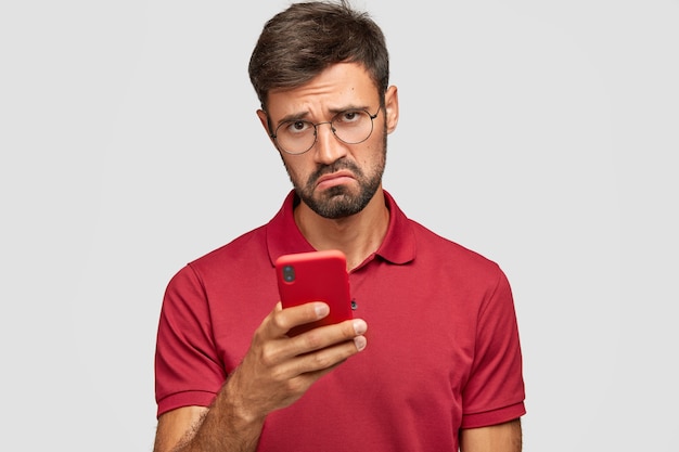 La foto di un giovane ragazzo con la barba lunga dispiaciuto tiene in mano un moderno smart phone rosso, insoddisfatto di leggere notizie negative su Internet, messaggi con gli amici nei social network, connesso a Internet wireless