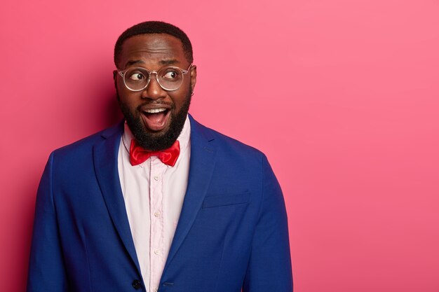 La foto di un uomo afroamericano con la barba lunga perplesso sorpreso tiene la bocca aperta, guarda con eccitazione da qualche parte, indossa un abito formale con papillon rosso