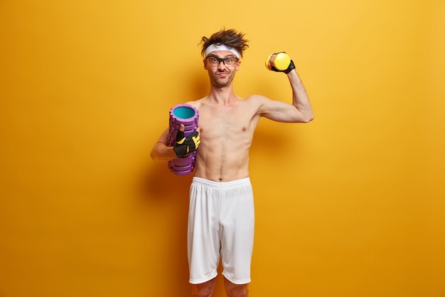 La foto di un uomo atletico determinato solleva il manubrio, posa con il rullo di schiuma, ha lo scopo di essere forte, gode di esercizi in palestra, isolato sul muro giallo. Persone, salute e concetto di fitness. Uno stile di vita sano