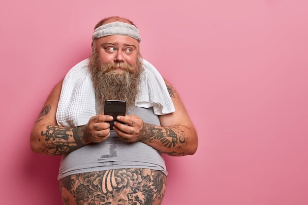 La foto di un uomo barbuto in sovrappeso legge sms sullo smartphone, impegnato a fare fitness a casa, controlla i risultati nell'app per lo sport quante calorie ha bruciato, ha la pancia tatuata che sporge da una maglietta sottodimensionata