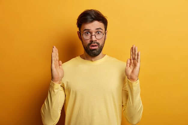 La foto di un uomo barbuto sbalordito solleva entrambi i palmi, forma qualcosa di molto grande e largo, eccitato per le dimensioni enormi, misura oggetti enormi, indossa occhiali trasparenti e un maglione giallo pastello casual. Troppo