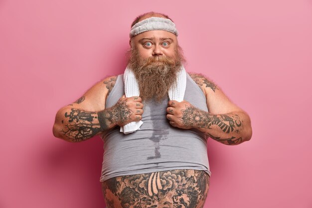 La foto di un uomo robusto e serio in abiti sportivi sogna un corpo muscoloso, lavora duramente sul suo corpo, vuole perdere peso, ha braccia tatuate, grande pancia, fa esercizi fisici con istruttore di fitness