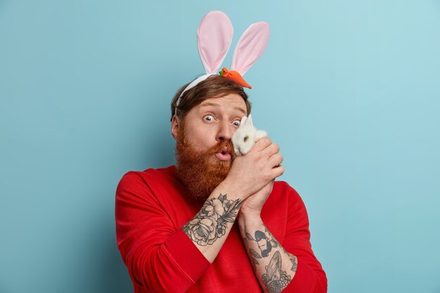 La foto di un uomo rosso sorpreso tiene un piccolo animale vicino al viso, gioca con il coniglio bianco, indossa orecchie da coniglio, si prepara per la festa in costume alla vigilia di Pasqua, posa contro il muro blu. Vacanze di primavera