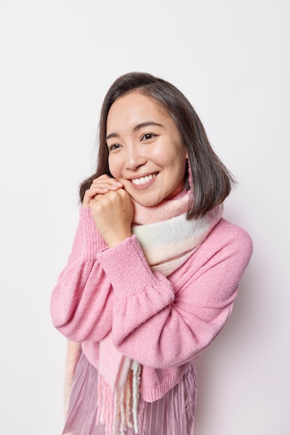 La foto di una bella donna asiatica sorride delicatamente tiene le mani vicino al viso ha un'espressione sognante pensa a qualcosa di piacevole indossa un maglione rosa e una sciarpa intorno al collo isolato su sfondo bianco