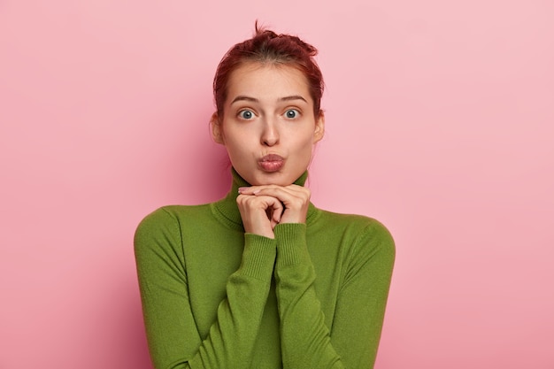 La foto di una bella donna europea mette il broncio sulle labbra, indossa un dolcevita verde, non ha trucco, tiene entrambe le mani sotto il mento, fa una smorfia alla telecamera, isolata sul muro rosa dello studio.