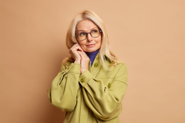 La foto di una bella donna rugosa di cinquant'anni tiene le mani vicino al viso e guarda da parte pensierosa vestita con un cappotto alla moda.