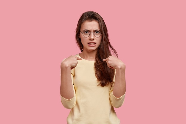 La foto di una donna adorabile imbarazzata indica se stessa, ha un'espressione del viso infastidita, indossa occhiali trasparenti e maglione, reagisce emotivamente quando viene incolpata, si trova al coperto sopra un muro rosa.