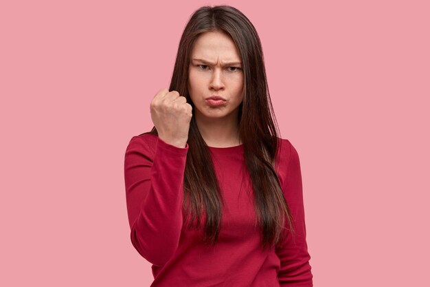 La foto di una donna arrabbiata aggrotta le sopracciglia, mostra il pugno, ha un'espressione del viso insoddisfatta, indossa abiti rossi casual, ha lunghi capelli lisci, minacce di qualcosa