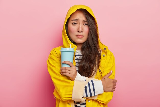 La foto di una donna asiatica insoddisfatta sorride, tiene le braccia incrociate, trema per il freddo dopo aver camminato sotto la pioggia, indossa un impermeabile giallo, tiene un caffè da asporto, si riscalda con una bevanda calda
