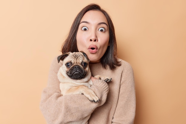 La foto di una donna asiatica scioccata fissa con gli occhi pieni di paura porta il cane carlino sulle mani non può credere in qualcosa di sorprendente indossa un maglione casual isolato su sfondo beige. Persone e animali domestici