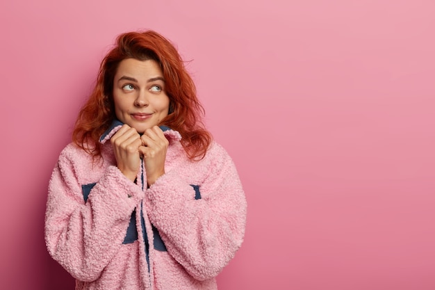 La foto di una donna attraente dai capelli rossi tiene le mani sotto il mento e guarda pensierosamente da parte, immagina di camminare all'aperto durante il tempo libero, indossa un cappotto invernale