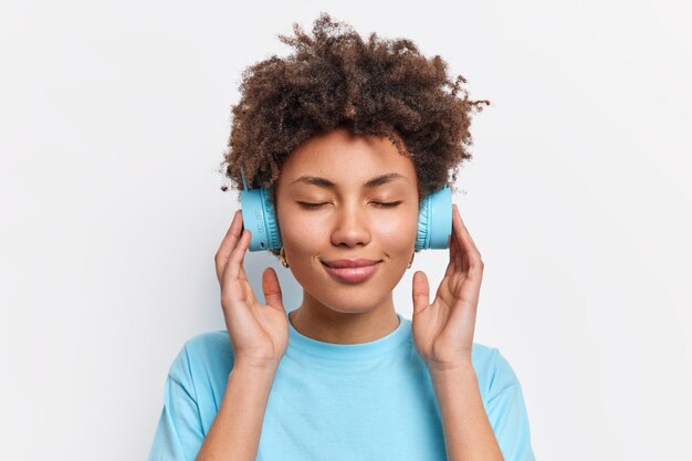 La foto di una donna dai capelli ricci rilassata chiude gli occhi e gode della musica tiene le mani sulle cuffie con una buona qualità del suono vestita con una maglietta blu casual isolata sul muro bianco. Concetto di stile di vita