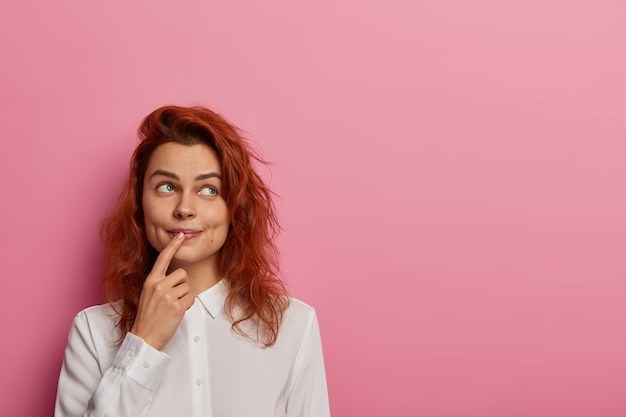 La foto di una donna dai capelli rossi sognante sogna ad occhi aperti qualcosa, guarda da parte, tiene il dito indice sulle labbra, indossa una camicia bianca