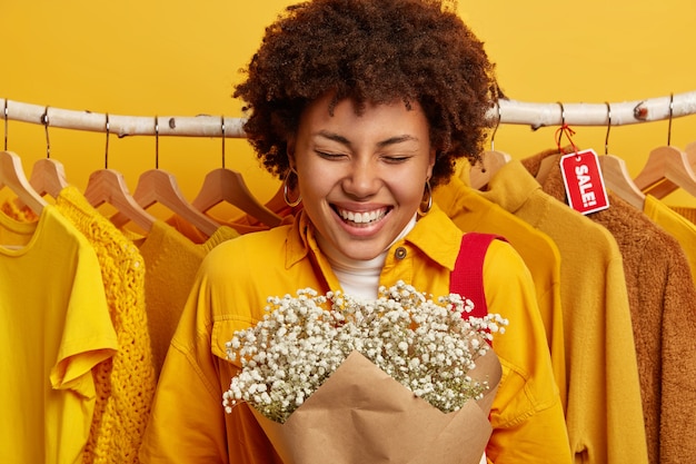 La foto di una donna felice tiene un bouquet, indossa un'elegante giacca gialla, sorride ampiamente, esulta, sta vicino ai vestiti sulle grucce