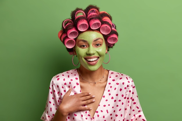 La foto di una donna gioiosa e spensierata si diverte a sottoporsi a procedure di bellezza a casa, indossa una maschera facciale verde per una pelle sana, indossa bigodini, indossa un abito di seta, sente qualcosa di divertente, posa al coperto