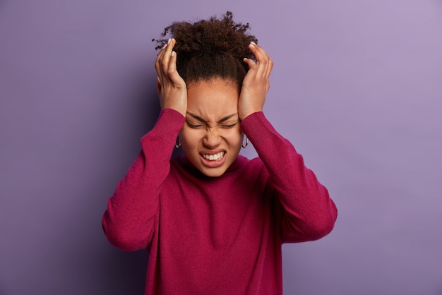 La foto di una donna insoddisfatta tiene le mani sulla tempia, soffre di un mal di testa insopportabile, stringe i denti per il dolore, è esausta dopo il lavoro, indossa un dolcevita bordeaux, isolato su un muro viola.