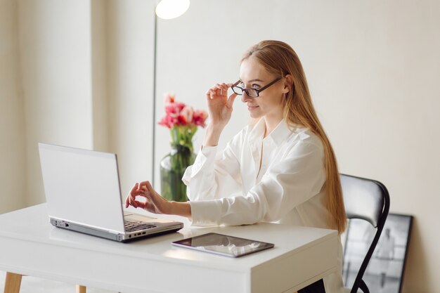 La foto di una giovane donna bionda allegra di affari in ufficio al chiuso lavora con il computer portatile e il telefono cellulare.