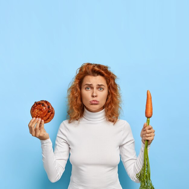 La foto di una giovane donna frustrata e delusa continua a dieta, porta il labbro inferiore, fa una scelta difficile tra panino e carota, un'alimentazione sana e cibo spazzatura, ha i capelli rossi ricci, un aspetto attraente