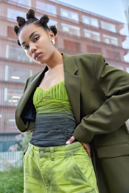 La foto di una giovane donna seria con l'acconciatura da panino e il trucco dell'eyeliner vestito con un abito alla moda tiene le mani sulla vita pone all'aperto sullo sfondo urbano focalizzato sulla fotocamera. Vista dal basso