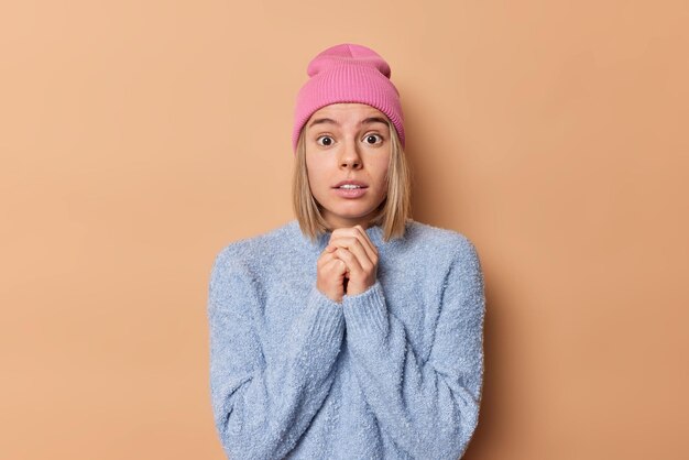 La foto di una giovane donna spaventata e preoccupata tiene le mani unite rimane senza parole sente grandi notizie non può credere ai propri occhi vestiti con abiti casual isolati su sfondo marrone. Reazioni umane.