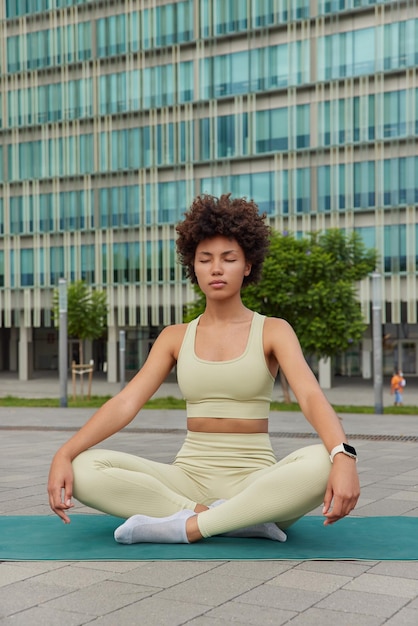 La foto di una giovane donna sportiva rilassata si siede nella posa del loto sul tappetino fitness indossa un top corto e leggings contro il grattacielo cerca di rilassarsi respira profondamente Meditazione della gente e stile di vita sano