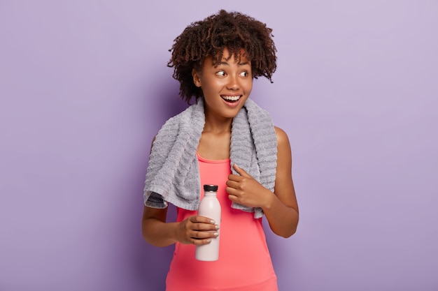 La foto di una sportiva dalla pelle scura deliziata ha un taglio di capelli afro, guarda da parte con un sorriso, vestita con un top rosa, porta una bottiglia, beve acqua mentre ha sete durante l'allenamento, si esercita al coperto.