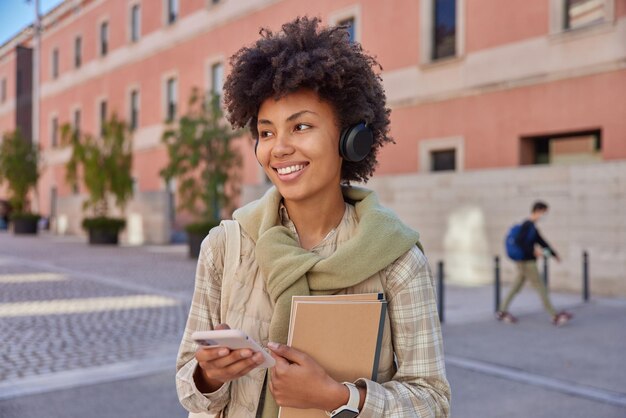 La foto di una studentessa allegra ascolta le informazioni tramite le cuffie stereo utilizza lo smartphone porta i blocchi note distoglie lo sguardo passeggia con gioia all'aperto sorride felicemente Concetto di persone e stile di vita