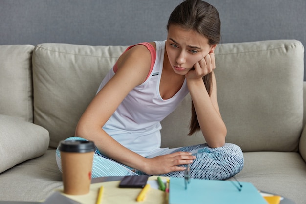 La foto di una studentessa stressata e sconvolta si sente stanca e guarda disperatamente in basso, si siede al desktop con carte e penne, t